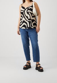 Schwarzes und beiges, gemustertes Camisole mit verstellbaren Trägern, kombiniert mit blauen Jeans mit geradem Bein und schwarzen klobigen Sandalen. Minimalistische weiße Tasche.