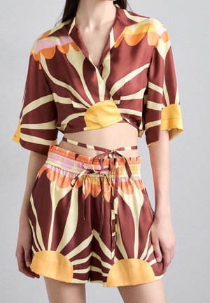 Kvinde iført todelt sæt med brun, gul, orange og lyserød abstrakt solstrålemønster, bestående af omslags-crop top og elastiske shorts i taljen.