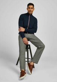 Mann, der auf einem schwarzen Hocker sitzt, trägt einen marineblauen Reißverschluss-Pullover, graue Hose und braune Sneaker vor einem schlichten grauen Hintergrund.