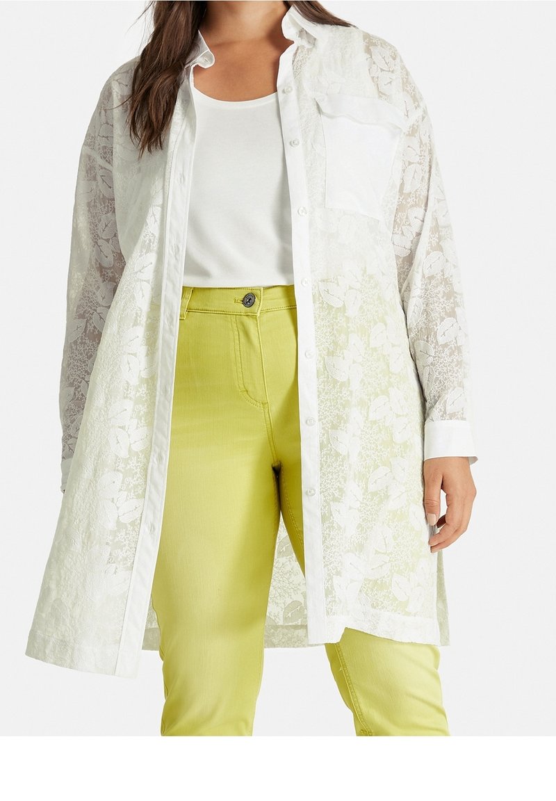 Femme portant une longue chemise blanche en dentelle florale par-dessus un top blanc et un pantalon jaune, vue du menton aux cuisses sur fond blanc.