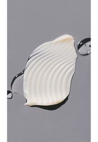 Vit, krämig kosmetisk produkt med texturerade vågor, placerad på en grå bakgrund. En droppe vätska ligger intill produkten.