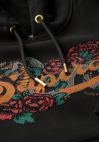 Czarna bluza z kapturem z kolorowym, wyszywanym wzorem kwiatowym i napisem "Superdry". Posiada złote elementy na sznurówkach oraz miękką teksturę.