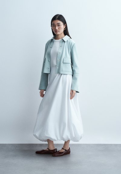 Γυναίκα που φοράει γαλάζιο κοντό σακάκι πάνω από λευκό φόρεμα μέχρι τον αστράγαλο με φουσκωτό τελείωμα και καφέ παπούτσια Mary Jane, στέκεται μπροστά σε απλό φόντο.