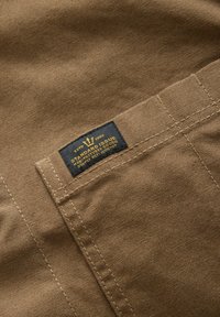 Brown fabric with a textured finish featuring a black label stitched onto a pocket, displaying yellow text and a crown logo.