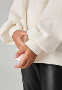 Felpa oversize color crema con polsini e orlo a coste, caratterizzata da una morbida texture del tessuto. Le mani sono unite sotto le maniche.