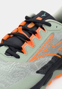 Lys grønne treningssko med svarte snører og oransje detaljer, med en teksturert overdel og en distinkt logo på tungen.