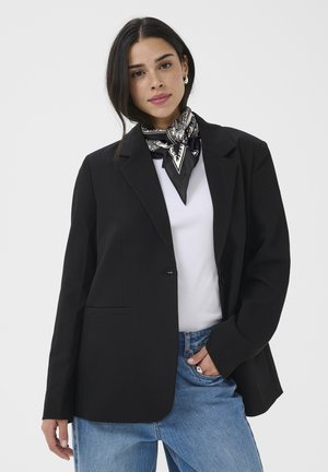 Žena v čiernom blejzeri, bielom topu, modrých džínsoch a čierno-bielom šatke na krk na jednotnom pozadí.