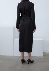 Robe noire à manches longues avec ceinture, en tissu lisse et brillant ; longueur genou, présentant un col classique et un design minimaliste.