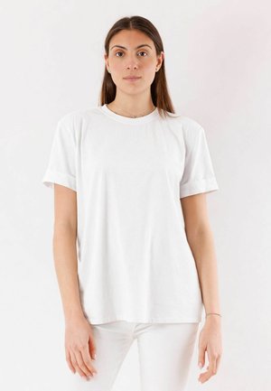 Giovane donna con capelli castani lisci, che indossa una maglietta bianca semplice e pantaloni bianchi, in piedi contro uno sfondo bianco.