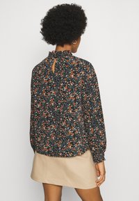 Svart blommig blus med långa ärmar, en volangkrage och en nyckelhålöppning bak, prydd med små orange och vita blommor.