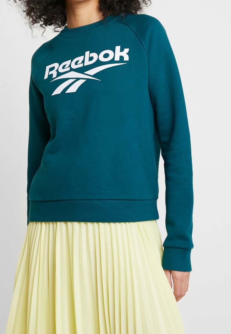 Femme portant un sweat-shirt Reebok bleu sarcelle et une jupe plissée jaune pâle sur un fond blanc uni.