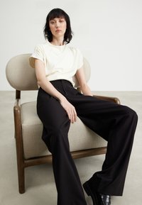 Top corto color crema a maniche corte abbinato a pantaloni neri a vita alta e gamba larga, seduto su una sedia in tessuto marrone chiaro.