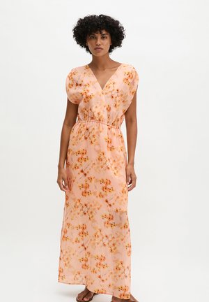 Femme portant une robe maxi sans manches florale pêche avec un décolleté en V et une taille froncée, se tenant devant un fond clair uni.