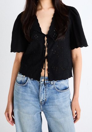 Mujer con blusa negra de manga corta con lazos delanteros y patrones florales recortados, combinada con jeans de talle alto en azul claro.