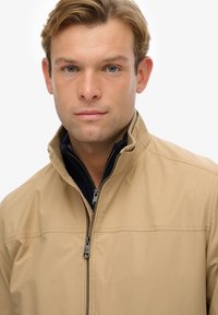 Lichtgewicht jack in tan met een hoge kraag en ritssluiting. Heeft een donkere binnenlaag die zichtbaar is bij de kraag en eenvoudige stikdetails.