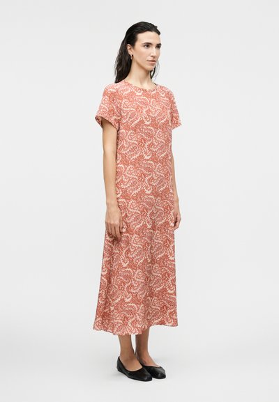 Γυναίκα που στέκεται φορώντας ένα κοραλλί φόρεμα κοντών μανικιών μέχρι τους αστραγάλους με λευκά λουλουδάτα σχέδια και μαύρα επίπεδα παπούτσια σε απλό φόντο.