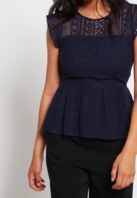 Top peplum blu navy con sovrapposizione di pizzo, maniche corte a cappuccio e intricati motivi a intaglio. Tessuto morbido e corpino aderente con orlo svasato.