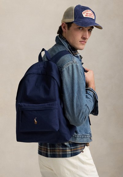 Jauns vīrietis, kurš valkā zilu džinsa jaku, rūtainu kreklu, tumši zilas krāsas mugursomu ar logotipu un zilu un bēšu cepuri, skatās pāri plecam.