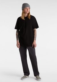 T-shirt noir oversize à manches courtes, associé à un pantalon gris et des chaussures slip-on à carreaux. Les accessoires comprennent un bonnet en tricot gris.