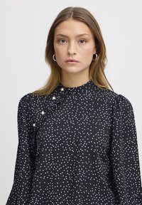 Blusa nera con pois bianchi, collo alto, dettagli arruffati e accenti simili a perle sul davanti, con maniche lunghe.