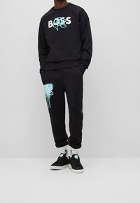 Sudadera negra y pantalones deportivos con texto blanco "BOSS" y detalles de diseño en turquesa. El material parece suave; ajuste casual con bolsillos. Zapatillas negras.