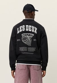 Μαύρο φούτερ με την επιγραφή "LES DEUX" σε έντονα λευκά γράμματα στην πλάτη, με γραφικό στοιχείο μπάσκετ και λεπτομέρειες κειμένου. Ριμπ μανσέτες και τελείωμα.