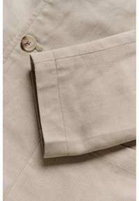 Beige fabric with a smooth texture, featuring a button closure and a folded sleeve. The stitching is neat with a herringbone pattern visible.