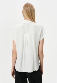Blusa blanca de manga corta con una textura suave, que presenta una costura en el yugo trasero y un dobladillo ligeramente curvado. Ligera y de corte relajado.