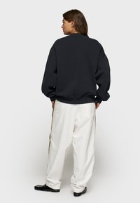 Felpa oversize grigio scuro con polsini a coste, abbinata a pantaloni bianchi larghi con strisce laterali. Scarpe nere basse completano l'outfit.
