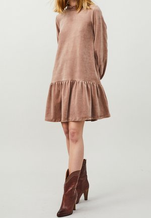 Femme portant une robe en velours rose poussiére à manches longues avec un ourlet à volants, associée à des bottes à talons en cuir marron mi-mollet, debout devant un fond uni.