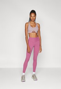 Helles lila Sport-BH mit einem Ausschnitt-Design, kombiniert mit hoch taillierten, schmal zulaufenden rosa Leggings. Die Schuhe sind hellgrau mit lila Akzenten.