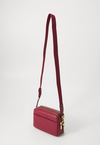 Raudona odinė crossbody rankinė su tekstūruota paviršiumi, dvigubais užtrauktukais ir auksinės spalvos detalėmis. Yra ilgas, reguliuojamas audinio diržas.