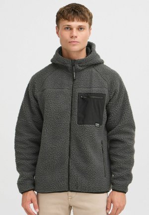 Veste en polaire grise avec une finition texturée, fermeture éclair à l'avant et poche poitrine noire. Elle possède une capuche et des poches latérales avec des poignets élastiques.