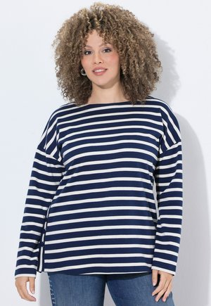 Kvinde med krøllet hår iført en langærmet top med navy og hvide striber og blå jeans, står foran en hvid baggrund.