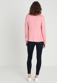Blazer rosa con maniche lunghe, tessuto testurizzato, design aderente e apertura frontale. Indossato con jeans skinny scuri e sneakers bianche.
