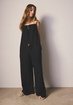 Kvinde med langt hår iført en løs sort jumpsuit med tynde stropper og perlebesat bindebånd, stående mod en lys tekstureret væg.