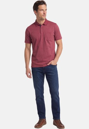 Mand iført en rød polo med korte ærmer, mørkeblå jeans og brune sneakers, stående med den ene hånd i lommen og kiggende til siden.