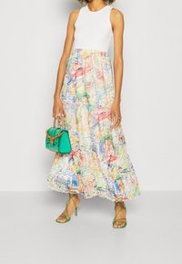Robe maxi colorée avec un corsage ajusté blanc et une jupe à volants en étages présentant un imprimé graphique. Les accessoires comprennent un petit sac à main vert.