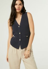 Tamnavija pletena prsluk s dubokim V-izrezom, s zlatnim gumbima i dva prednja džepa. Nositi preko svijetlosmeđih hlača.