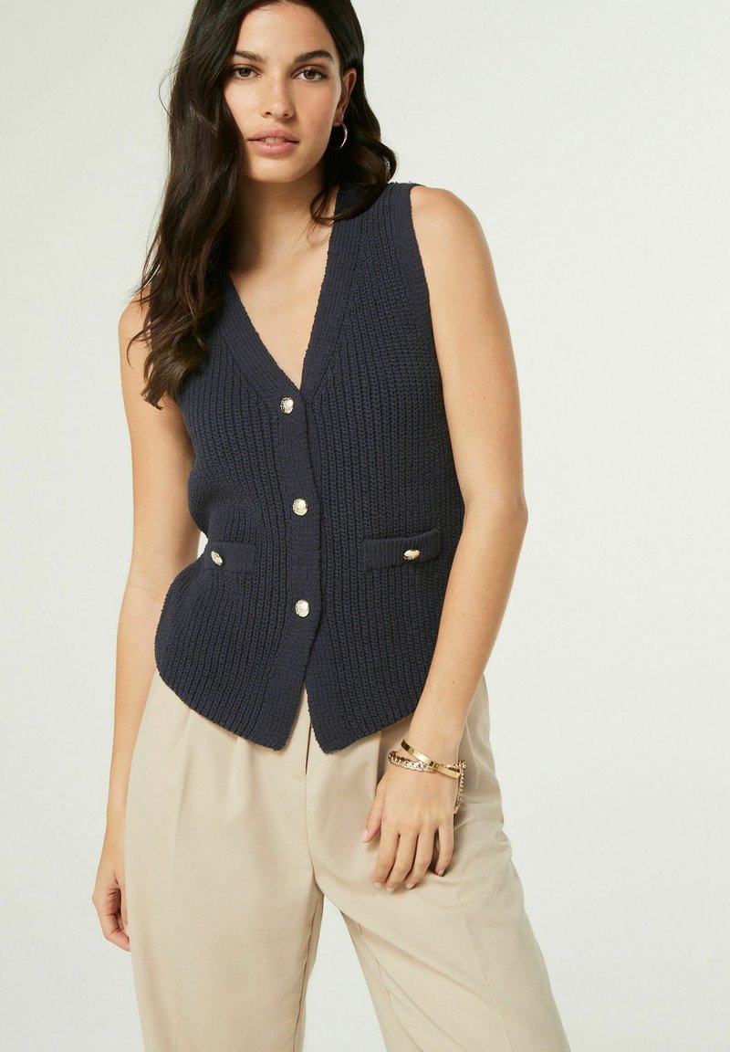 Tamnavija pletena prsluk s dubokim V-izrezom, s zlatnim gumbima i dva prednja džepa. Nositi preko svijetlosmeđih hlača.