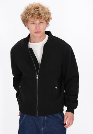 Ung mand med krøllet blond hår iført en sort zip-up jakke over en hvid skjorte og blå jeans, stående foran en hvid baggrund.