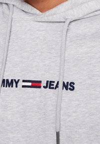 Szara bluza z kapturem z granatowym haftowanym logo "TOMMY JEANS" oraz poziomym emblematem w czerwone, białe i czarne paski.