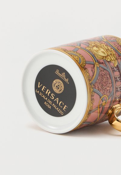 Λευκή και ροζ πορσελάνινη κούπα Versace με περίτεχνα χρυσά και ανθικά σχέδια, χρυσή λαβή, μαύρη βάση με ετικέτα που δείχνει το εμπορικό σήμα και το όνομα του μοτίβου.