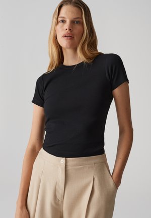 Schwarzes, geripptes T-Shirt mit Kurzarm aus modalisierendem Material, kombiniert mit beigen, hochtaillierten, plissierten Hosen mit Knopfverschluss.