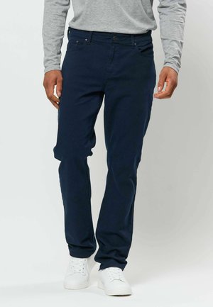 Homme portant un pantalon bleu foncé, une chemise grise à manches longues et des baskets blanches, debout devant un fond clair uni.