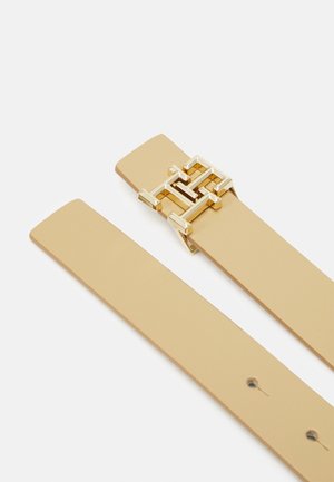 Ceinture en cuir beige avec une boucle rectangulaire dorée brillante présentant un motif géométrique enchevêtré, présentée à plat sur un fond blanc.
