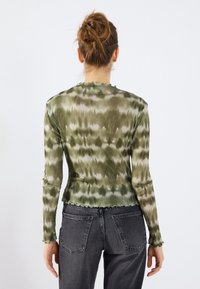 Long-sleeve green tie-dye top with a textured finish and ruffled hem, paired with high-waisted gray jeans featuring visible stitching details.