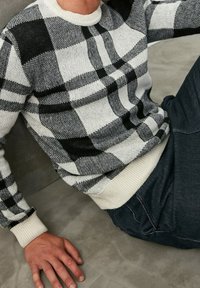 Jersey de punto en patrón de cuadros en blanco y negro, con cuello redondo acanalado y puños. Textura suave con cinturilla en contraste y detalles sutiles.