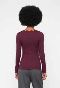 Žena s afro účesom, oblečená v priliehavom dlhom rukáve burgundskej blúzke s čipkovými detailmi na manžetách a výstrihu, v kombinácii so šedými nohavicami.