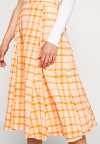 Pleated, puolipitkä hame oranssissa ja persikanvärisessä ruutukuosissa. Valmistettu kevyestä kankaasta, jossa on sileä pinta ja istuva vyötärö.